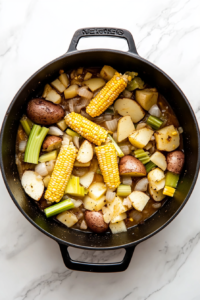 This image shows fresh corn kernels and juices from the cobs being stirred into a mixture of vegetables in a Dutch oven, intensifying the sweet and savory flavors of the chowder.