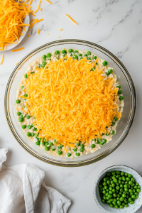 This image shows shredded cheese being sprinkled generously over the peas layer, adding a creamy and rich element to the layered salad.