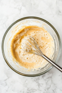 This image shows a hand whisking mashed bananas, eggs, and other wet ingredients together in a clear glass mixing bowl, creating a smooth and well-blended mixture for banana bread.