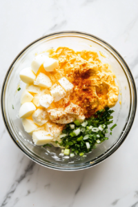 This image shows paprika, salt, and black pepper being sprinkled over the egg salad mixture inside a bowl, enhancing the flavor and color.