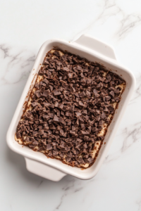 This image shows chocolate toffee bits being evenly scattered over a layer of pressed cake mix dough inside a ceramic baking dish. The setup is clean and minimal, with the dish resting on a white marble countertop.