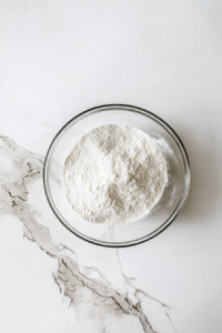 This image shows sifted flour, baking powder, and salt in a glass bowl on a white marble countertop.