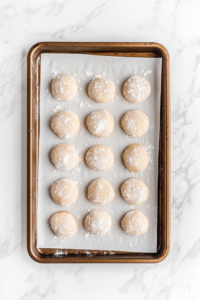 This image shows milk cookie dough rounds on a baking tray lightly sprinkled with granulated sugar, resting on a clean white marble countertop.